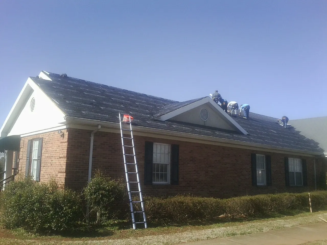 Crew tearing shingles off a brick ranch roof with a ladder set — Emergency Roof Repair work by our crew in Mount Pleasant