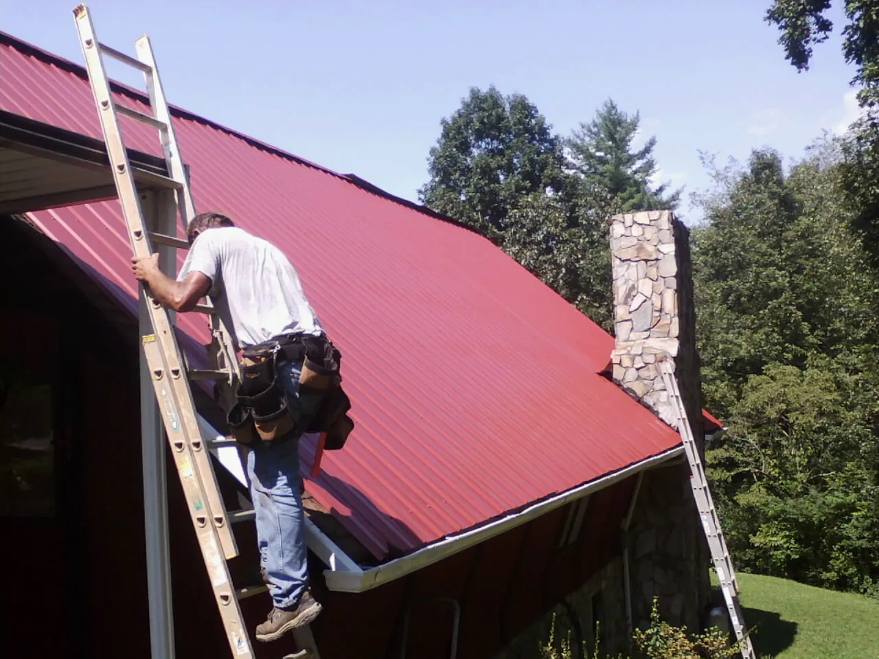 Roofer on a ladder beside a red metal roof and stone chimney — Soffit & Fascia Repair by Roofing Brothers in Mount Pleasant